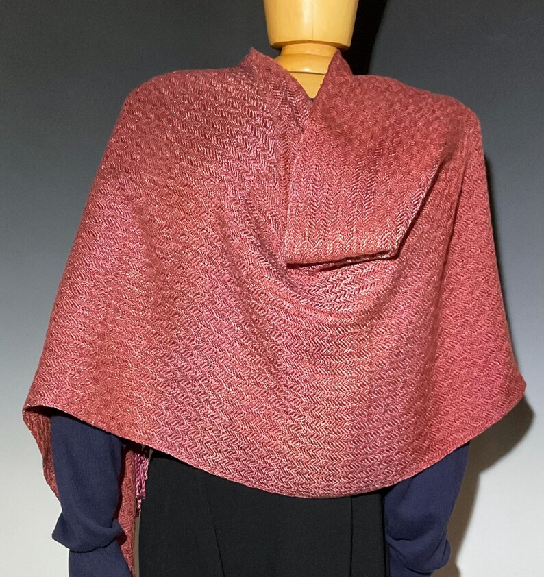 May include: A woven shawl in shades of red and pink, draped over a mannequin. The shawl features a textured, herringbone pattern. The mannequin is wearing a dark blue long-sleeved shirt and a black skirt.