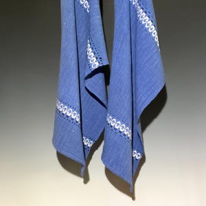 May include: A blue textile hanging vertically, featuring a woven texture and a horizontal band of white decorative embroidery with small black accents. The fabric displays a consistent weave pattern throughout, with clean edges and a soft drape.