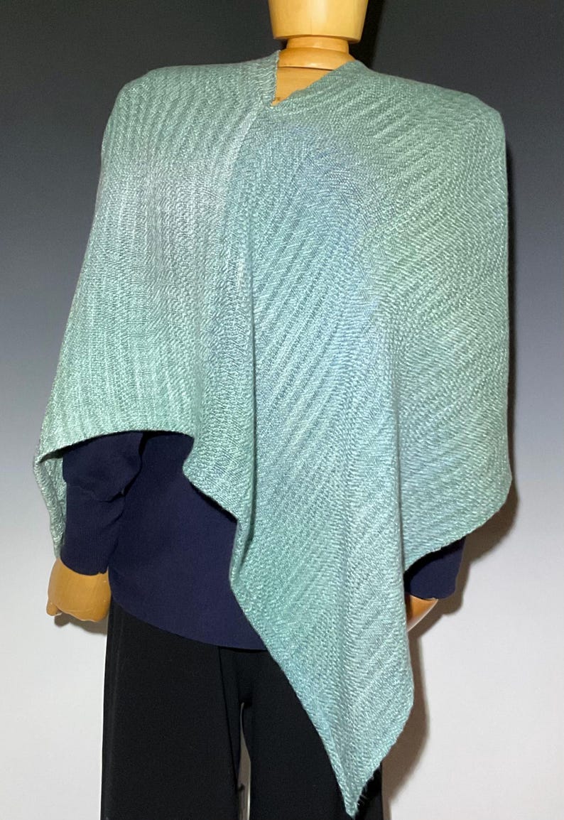 Pu&ograve; includere: Un poncho verde acqua chiaro con un motivo a trama diagonale strutturato. Il poncho &egrave; drappeggiato su un manichino, rivelando una camicia blu navy a maniche lunghe e pantaloni neri. L'indumento ha una vestibilit&agrave; rilassata e una sottile sfumatura di colore.