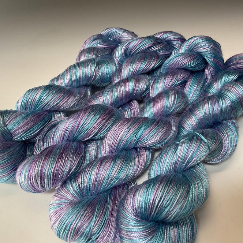 May include: A close-up of several skeins of yarn in shades of blue, teal, and purple. The yarn has a soft, silky appearance, with subtle variations in color throughout each strand. The yarn is likely used for knitting or crocheting projects.