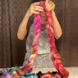 May include: A person holding two long, braided strands of hand-dyed yarn. The yarn transitions from pink to red hues. Other skeins of yarn in various colors are visible below, suggesting a craft or textile theme.