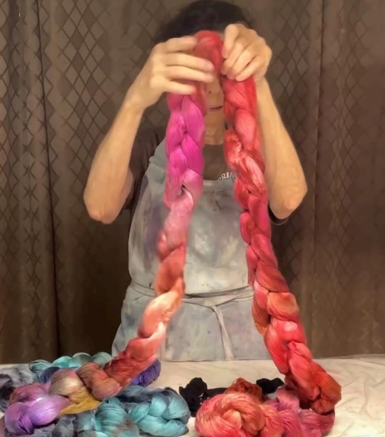 May include: A person holding two long, braided strands of hand-dyed yarn. The yarn transitions from pink to red hues. Other skeins of yarn in various colors are visible below, suggesting a craft or textile theme.