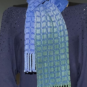 May include: A woven scarf in shades of blue and green. The scarf features a geometric pattern and fringe at the ends. The scarf is draped over a dark blue top.