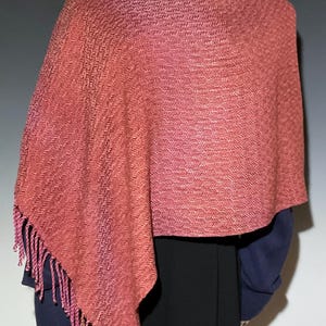 May include: A woven shawl in shades of coral and pink, draped over a mannequin. The shawl features a textured pattern and fringed edges. The garment is a wrap, suitable for cool weather.