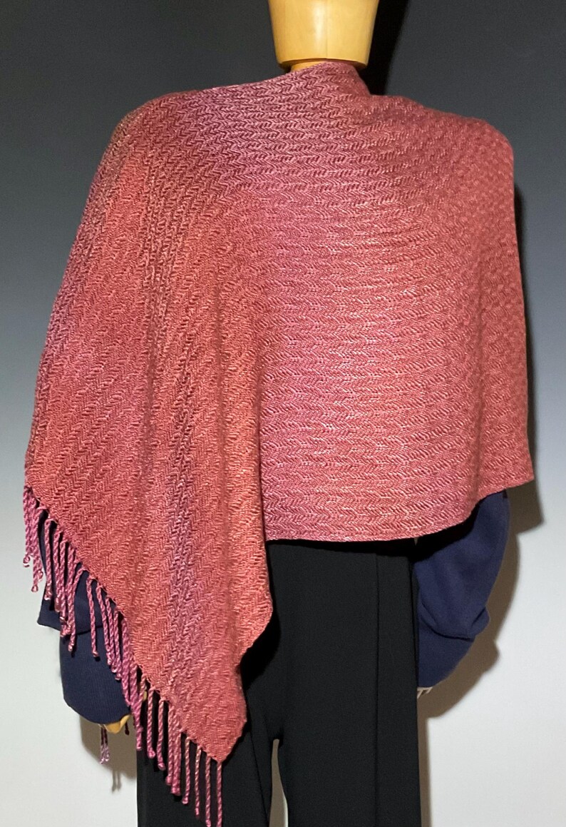 May include: A woven shawl in shades of coral and pink, draped over a mannequin. The shawl features a textured pattern and fringed edges. The garment is a wrap, suitable for cool weather.