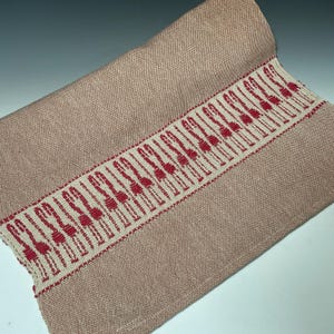 Puede incluir: Textil tejido enrollado, de color rosa apagado. Una banda decorativa de tela blanquecina con un patr&oacute;n rojo repetido recorre un borde. El textil parece ser un camino de mesa o un art&iacute;culo de decoraci&oacute;n del hogar similar.