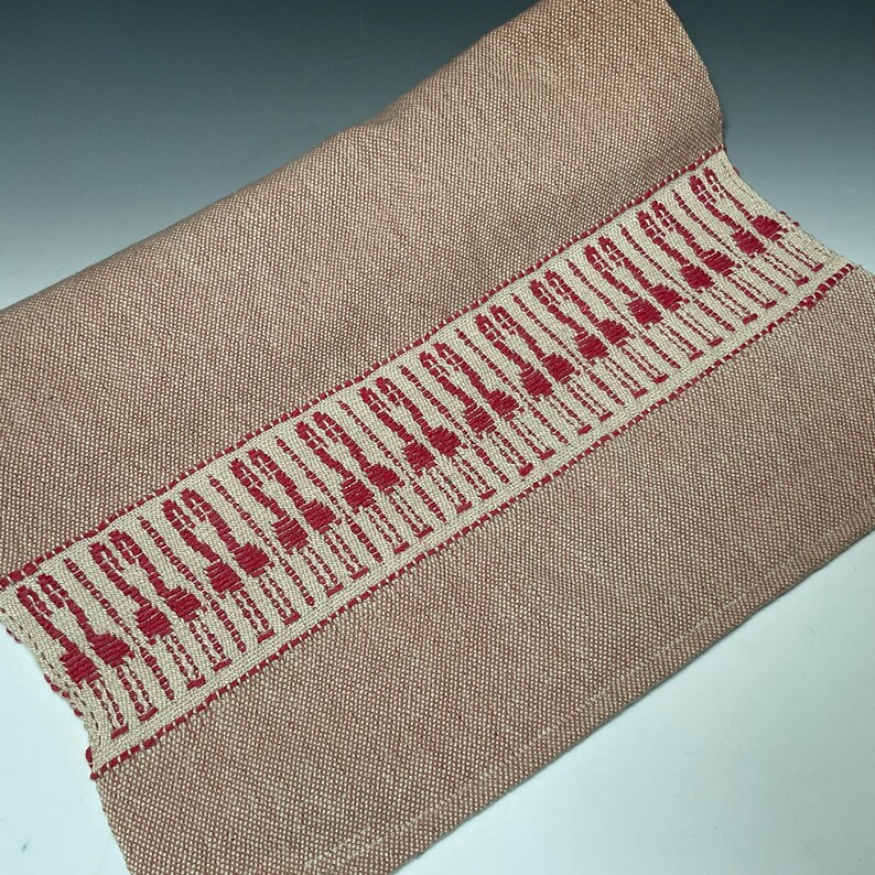 Puede incluir: Textil tejido enrollado, de color rosa apagado. Una banda decorativa de tela blanquecina con un patr&oacute;n rojo repetido recorre un borde. El textil parece ser un camino de mesa o un art&iacute;culo de decoraci&oacute;n del hogar similar.