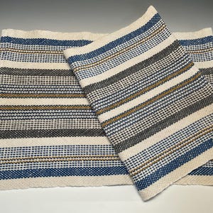 May include: Handwoven rectangular placemat and matching napkin. The off-white fabric features horizontal stripes in navy blue, gray, and gold. The placemat is rectangular, and the napkin is folded and placed on top. The weave is visible.
