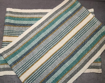 May include: Two woven rugs with a striped pattern in shades of gray, teal, and gold. The rugs are made of a natural fiber and have a textured surface.