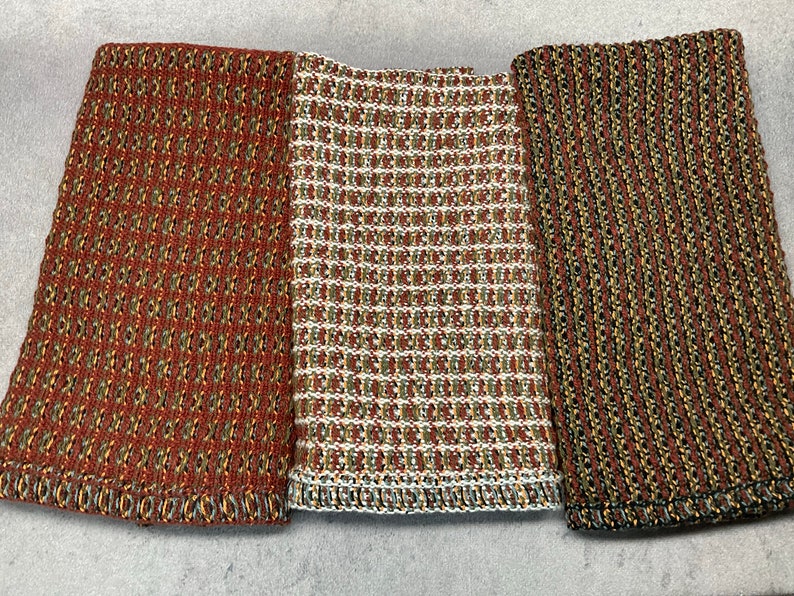 May include: Three kitchen towels with a woven texture. The towels are in shades of brown, green, and white. The towels are lined up side by side.