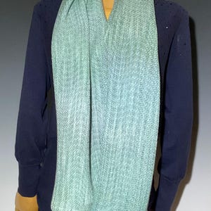 Pu&ograve; includere: Una sciarpa verde chiaro e blu drappeggiata su una maglia a maniche lunghe blu navy. La sciarpa ha una trama strutturata e un effetto di colore sfumato. La sciarpa &egrave; esposta su un manichino di legno.