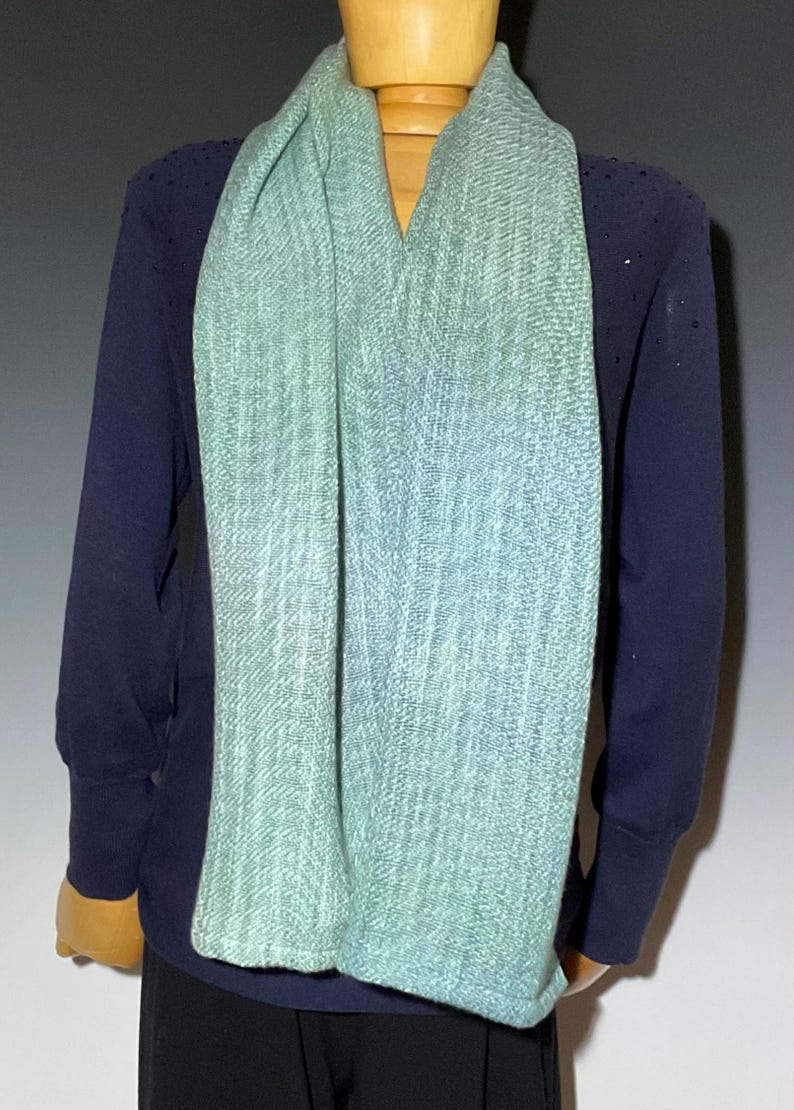 Pu&ograve; includere: Una sciarpa verde chiaro e blu drappeggiata su una maglia a maniche lunghe blu navy. La sciarpa ha una trama strutturata e un effetto di colore sfumato. La sciarpa &egrave; esposta su un manichino di legno.