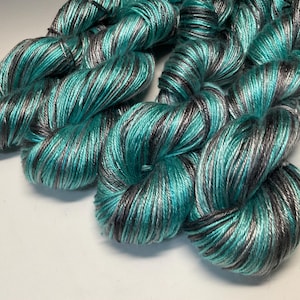 Op de afbeelding: Close-up van verschillende strengen garen in tinten van blauwgroen, grijs en zwart. Het garen is strak gewikkeld, met een lichte glans, en ziet er zacht uit. De kleurvariaties cre&euml;ren een textuureffect, geschikt voor brei- of haakprojecten.