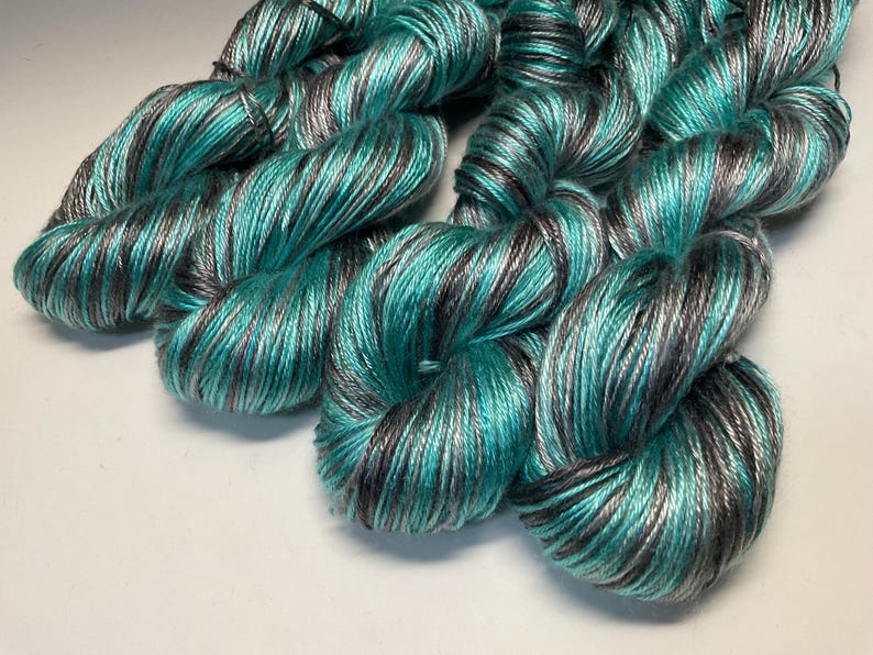 Op de afbeelding: Close-up van verschillende strengen garen in tinten van blauwgroen, grijs en zwart. Het garen is strak gewikkeld, met een lichte glans, en ziet er zacht uit. De kleurvariaties cre&euml;ren een textuureffect, geschikt voor brei- of haakprojecten.