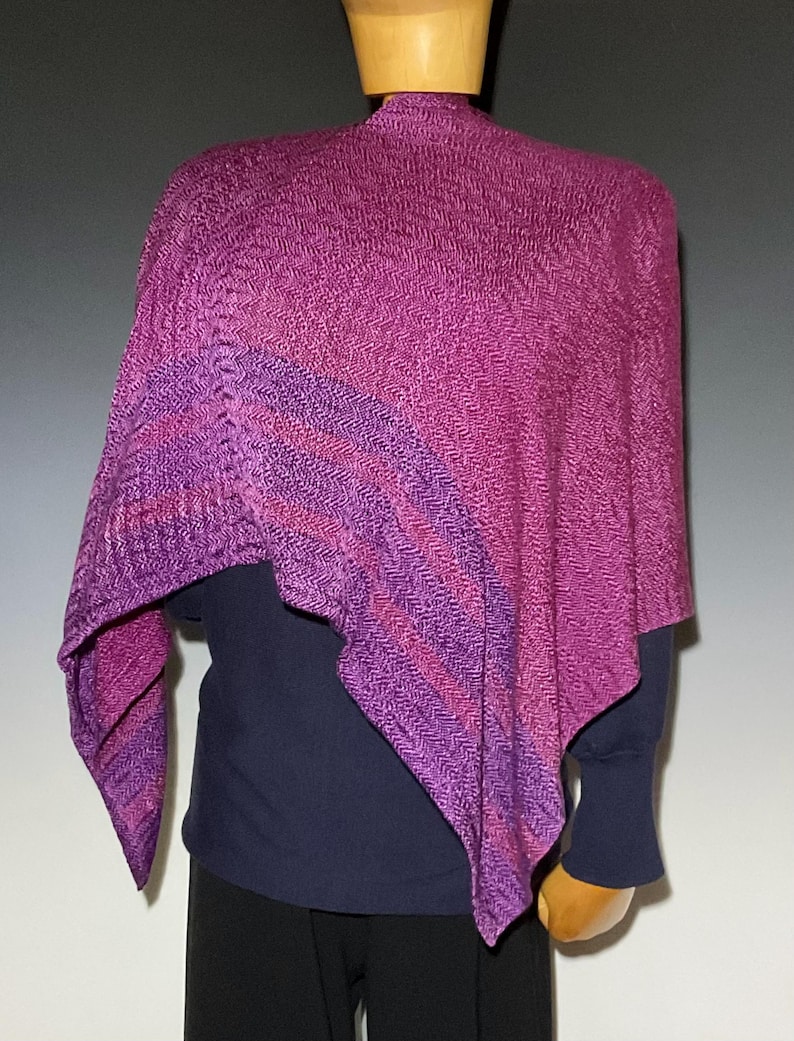 May include: A knitted poncho featuring a vibrant magenta and purple herringbone pattern. The garment has an asymmetrical drape with a textured surface and a deep, saturated color palette that transitions between shades of pink, violet, and purple.