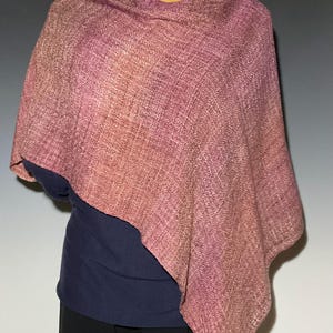 May include: A woven poncho featuring a gradient of dusty rose, mauve, and deep purple tones. The fabric displays a visible textured weave pattern with a soft drape, covering the upper torso and shoulders in an asymmetrical, loose-fitting silhouette.