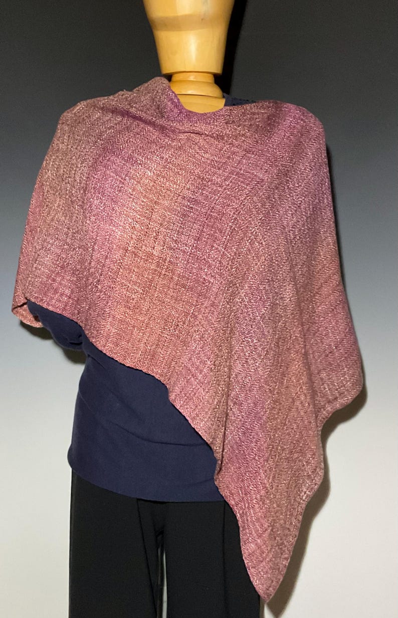 May include: A woven poncho featuring a gradient of dusty rose, mauve, and deep purple tones. The fabric displays a visible textured weave pattern with a soft drape, covering the upper torso and shoulders in an asymmetrical, loose-fitting silhouette.