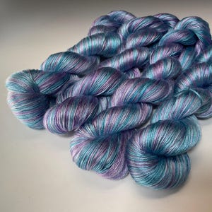 May include: Skeins of hand-dyed yarn in shades of teal, purple, and white. The yarn is arranged in a loose pile, with the individual strands visible. The colors blend together to create a soft, watercolor effect. Ideal for knitting, crocheting, or other fiber arts projects.