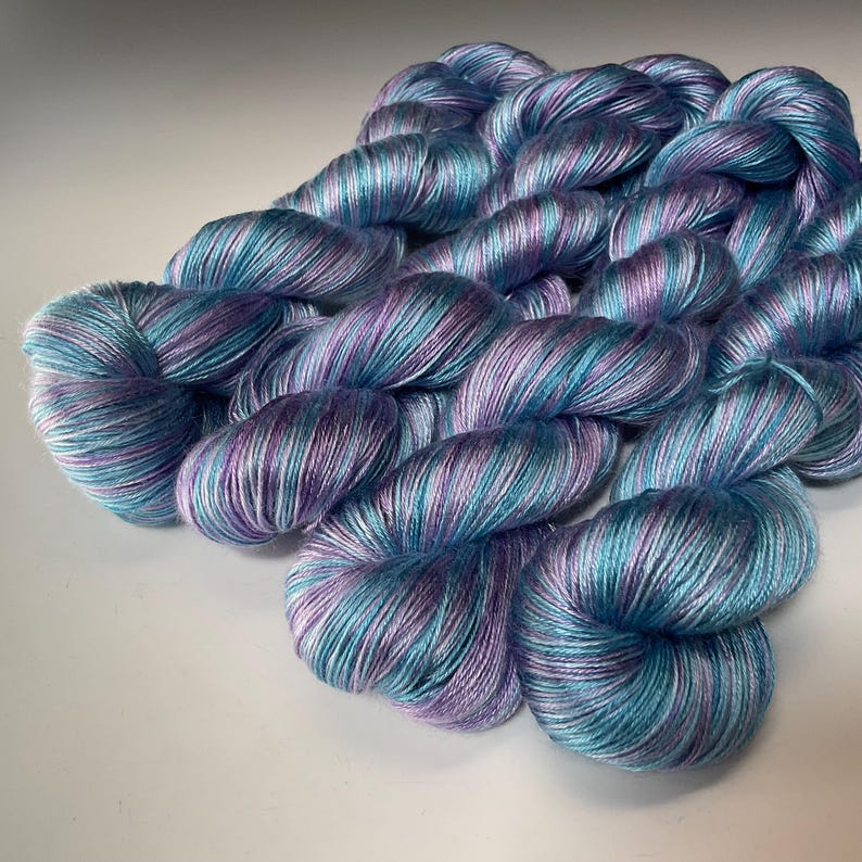 May include: Skeins of hand-dyed yarn in shades of teal, purple, and white. The yarn is arranged in a loose pile, with the individual strands visible. The colors blend together to create a soft, watercolor effect. Ideal for knitting, crocheting, or other fiber arts projects.