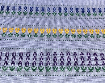 May include: A close-up of a woven fabric with a repeating pattern of purple, yellow, and green flowers. The fabric has a light blue background.