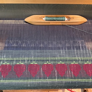 Puede incluir: Un telar con una urdimbre azul oscuro y una trama verde. El telar está configurado para tejer un patrón de corazones rojos.