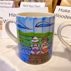 Könnte beinhalten: Eine weiße Keramiktasse mit einem blauen, grünen und braunen bemalten Design von zwei Personen in Badeanzügen, die auf einem Steg sitzen. Der Text "I LOVE THA" ist auf den Boden der Tasse gemalt.