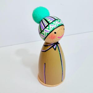 以下が含まれることがあります： 顔が描かれた木製のペグドール。緑色のポンポンが付いた模様入りの帽子をかぶっています。人形は、黒と紫でペイントされたディテールを持つ、タン色のボディをしています。人形の高さは約13cmです。