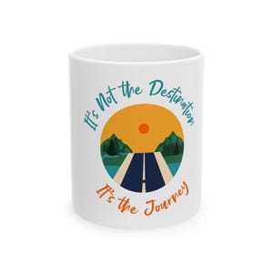 May include: White ceramic mug featuring a scenic design with a road leading towards a sunset. The design includes mountains, trees, and the text "It's Not the Destination, It's the Journey" in teal and orange.