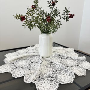 以下が含まれることがあります： 繊細な花柄の白いクロシェのテーブルランナー。テクスチャのある表面の白い花瓶には、赤い実のついた緑の枝のブーケが活けられています。