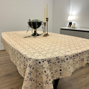 Könnte beinhalten: Eine beige Häkeltischdecke mit einem zarten Blumenmuster. Die Tischdecke liegt auf einem Holztisch mit einer schwarzen Schüssel und goldenen Kerzenständern.