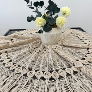 May include: A beige crocheted table runner with a delicate pattern of circles and scallops. The runner is displayed on a table with a white vase filled with yellow flowers and green leaves.
