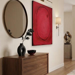 Op de afbeelding: Een houten dressoir met twee deuren, bekroond door een zwarte vaas met donker gebladerte, een kleine donkere kom, een ronde spiegel met een dunne lijst en een groot rechthoekig canvas met een rode achtergrond, zwarte ovale omtrek en rode cirkel.