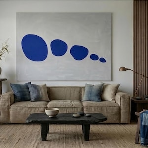 Peut inclure: Une grande œuvre d'art murale rectangulaire présentant cinq formes organiques bleu uni de tailles variées sur un fond gris clair, placée au-dessus d'un canapé en lin beige avec des coussins bleus et beiges et une table basse en bois noir.