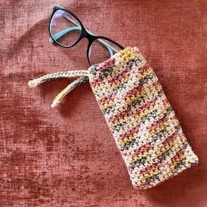Peut inclure: Des lunettes noires avec des accents turquoise sont placées dans un étui à lunettes en crochet fait à la main. L'étui présente un mélange de couleurs, notamment du rose, du jaune, du gris et de la crème. Il possède une fermeture à cordon.