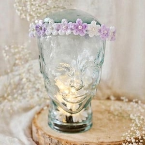Könnte beinhalten: Ein Blumenkranz mit gehäkelten weißen und lavendelfarbenen Blüten und Perlenakzenten. Der Kranz wird auf einem durchsichtigen Glaskopf auf einer Holzscheibe mit einem weichen, verschwommenen Hintergrund präsentiert.