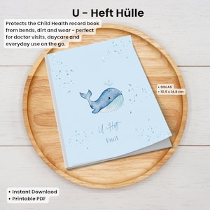 Könnte beinhalten: Hellblaue Umschlag für das Kinderuntersuchungsheft mit einer Walillustration. Der Umschlag trägt die Aufschrift "U - Heft Hülle" und den Text "DIN A6 - 10,5 x 14,8 cm". Das Buch liegt auf einem Holztablett.
