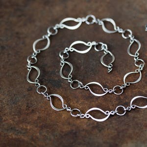 May include: A silver chain necklace with a unique, flowing design. The chain is made of interconnected, elongated, teardrop-shaped links.