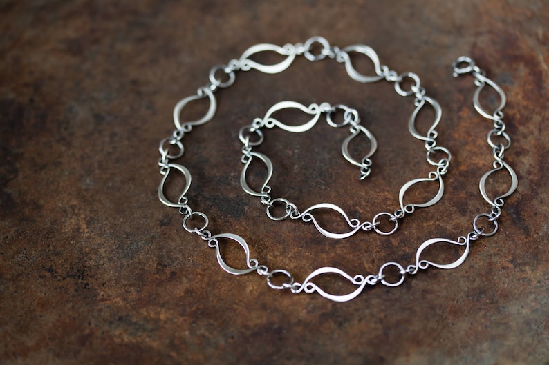 May include: A silver chain necklace with a unique, flowing design. The chain is made of interconnected, elongated, teardrop-shaped links.