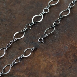 May include: A silver chain necklace with a delicate, open-link design. The links are shaped like elongated ovals with a slight twist. The chain is shown on a brown, textured surface.