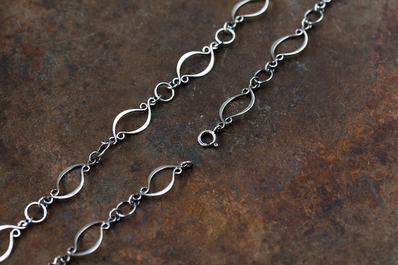 May include: A silver chain necklace with a delicate, open-link design. The links are shaped like elongated ovals with a slight twist. The chain is shown on a brown, textured surface.