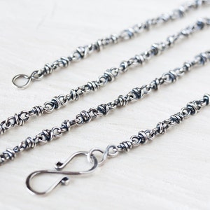 May include: A silver chain necklace with a twisted design. The chain is made of small, interconnected links that create a unique and intricate pattern. The necklace has a lobster clasp closure.