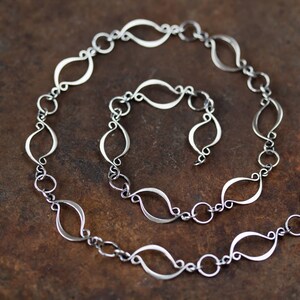May include: A silver chain necklace with a unique, delicate design featuring interconnected, elongated oval links. The chain is coiled on a brown surface.