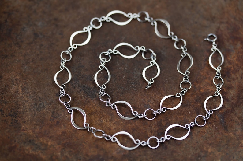 May include: A silver chain necklace with a unique, delicate design featuring interconnected, elongated oval links. The chain is coiled on a brown surface.