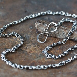 May include: A silver chain necklace featuring a distinctive twisted design and a simple hook clasp. The chain is crafted from small, interlinked loops, resulting in a textured appearance.