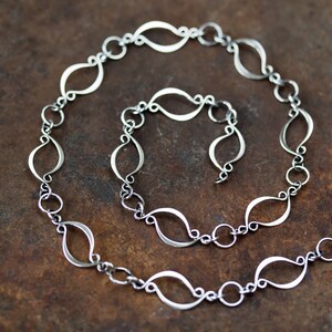 May include: A silver chain necklace with a unique, flowing design. Each link is shaped like a teardrop, creating a delicate and elegant look.