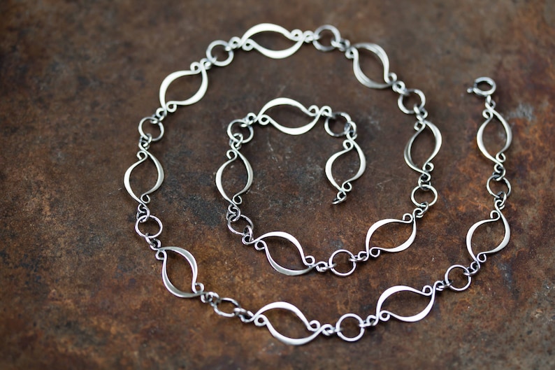 May include: A silver chain necklace with a unique, flowing design. Each link is shaped like a teardrop, creating a delicate and elegant look.