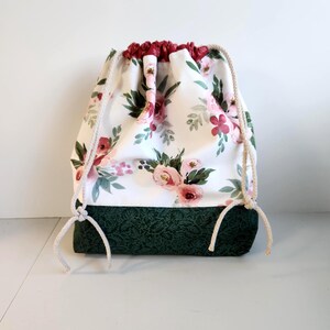 May include: A drawstring bag with a white floral pattern featuring pink roses and green leaves. The bottom of the bag is dark green, and the drawstring is a cream color. The top of the bag is a deep red.