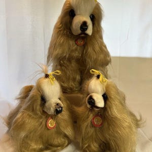 May include: Three plush Cocker Spaniel dog figures. The dogs have tan fur, white faces, and black noses. Each dog has a yellow bow in its hair and a red and gold medallion. The dogs are posed together.