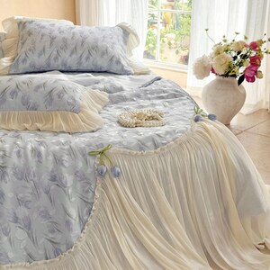 May include: A light blue bedding set featuring a floral tulip pattern, accented with cream-colored ruffled edges and decorative fabric tulip embellishments. The set includes a comforter and matching pillowcases with soft, layered fabric textures.