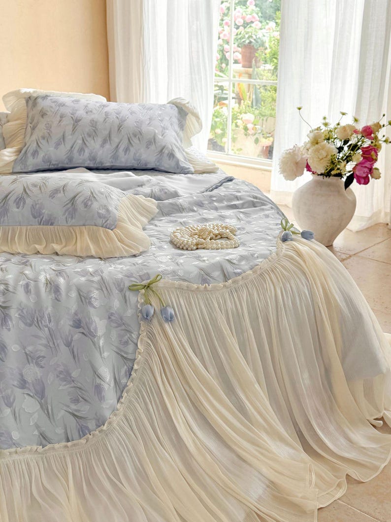 May include: A light blue bedding set featuring a floral tulip pattern, accented with cream-colored ruffled edges and decorative fabric tulip embellishments. The set includes a comforter and matching pillowcases with soft, layered fabric textures.